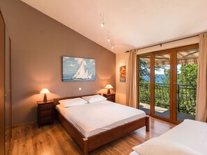 A bedroom with a wooden bed, two bedside lamps, a painting of sailboats, and a balcony with a view of greenery and the sea. - Irena Apartments 2 kat / HR-10649-02 by Interhome (Selce)