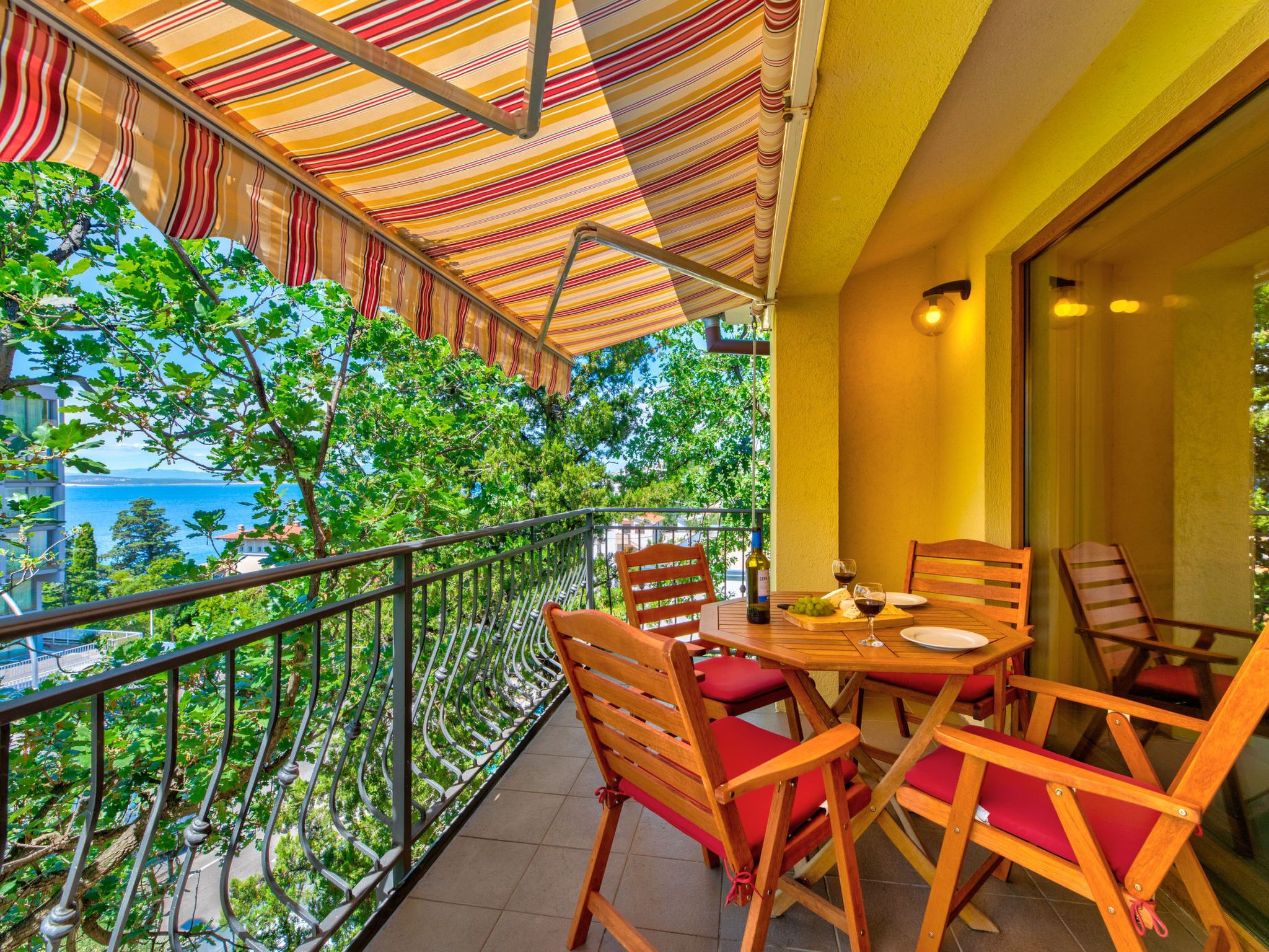 Un balcon agrémenté d’un auvent à rayures, de chaises en bois, d’une table avec une bouteille et des verres, et offrant une vue sur la mer et les bâtiments.