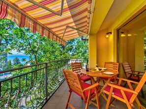 Un balcon agrémenté d’un auvent à rayures, de chaises en bois, d’une table avec une bouteille et des verres, et offrant une vue sur la mer et les bâtiments.