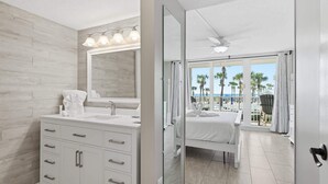 A modern bathroom with a large mirror, white vanity, and a view of a balcony with palm trees and a pool. - Dunes of Panama C104 (Panama City Beach)