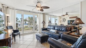 A spacious living room with a leather sofa, a coffee table, and a TV. There is a balcony with a view of palm trees and buildings. - Dunes of Panama C104 (Panama City Beach)
