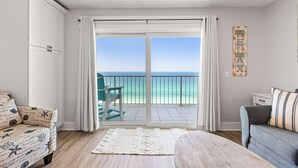 Quarto com uma grande porta de vidro deslizante que dá para uma varanda com vista para o oceano. Há uma cadeira verde e um sofá cinza com almofadas listradas.