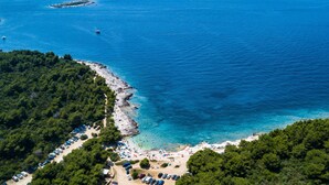Oversigtsbillede af et kystområde med en strand, klart blåt vand og en parkeringsplads med biler.