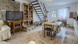 Een gezellige woonkamer met een televisie, houten meubels, een boekenkast, een eettafel met stoelen, een trap en een keukenhoek.