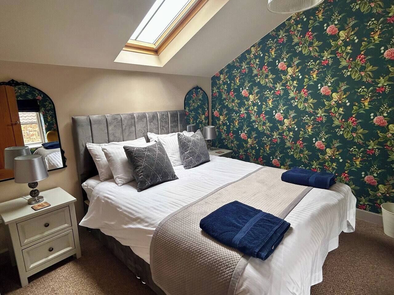 A bedroom with a bed, a nightstand, a lamp, a mirror, and a floral wallpaper.