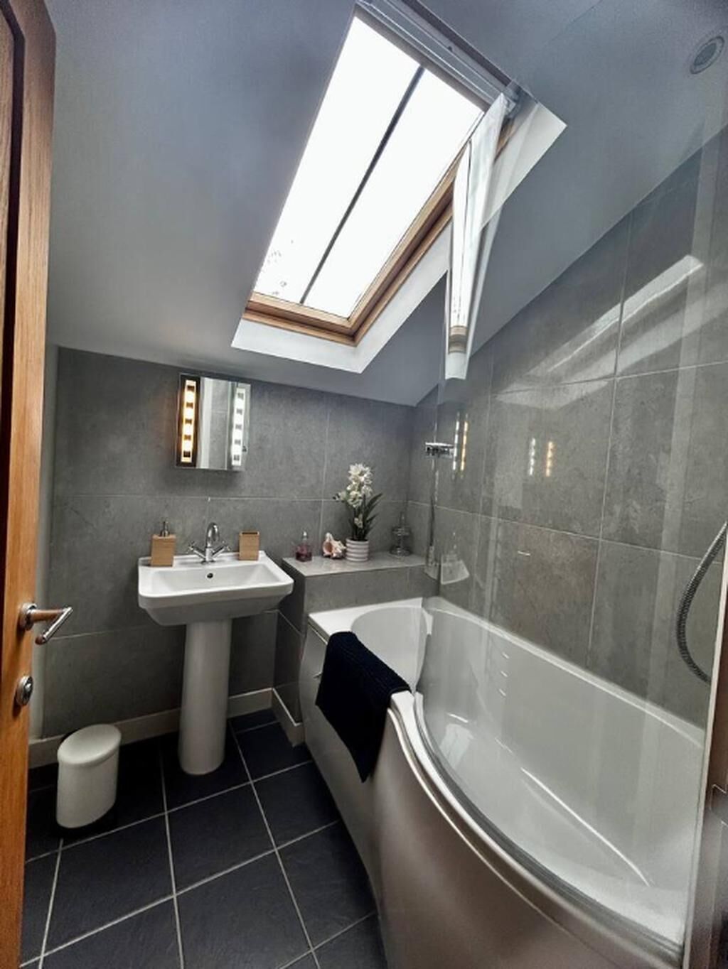 A modern bathroom with a skylight, a freestanding bathtub, a pedestal sink, and a small mirror.