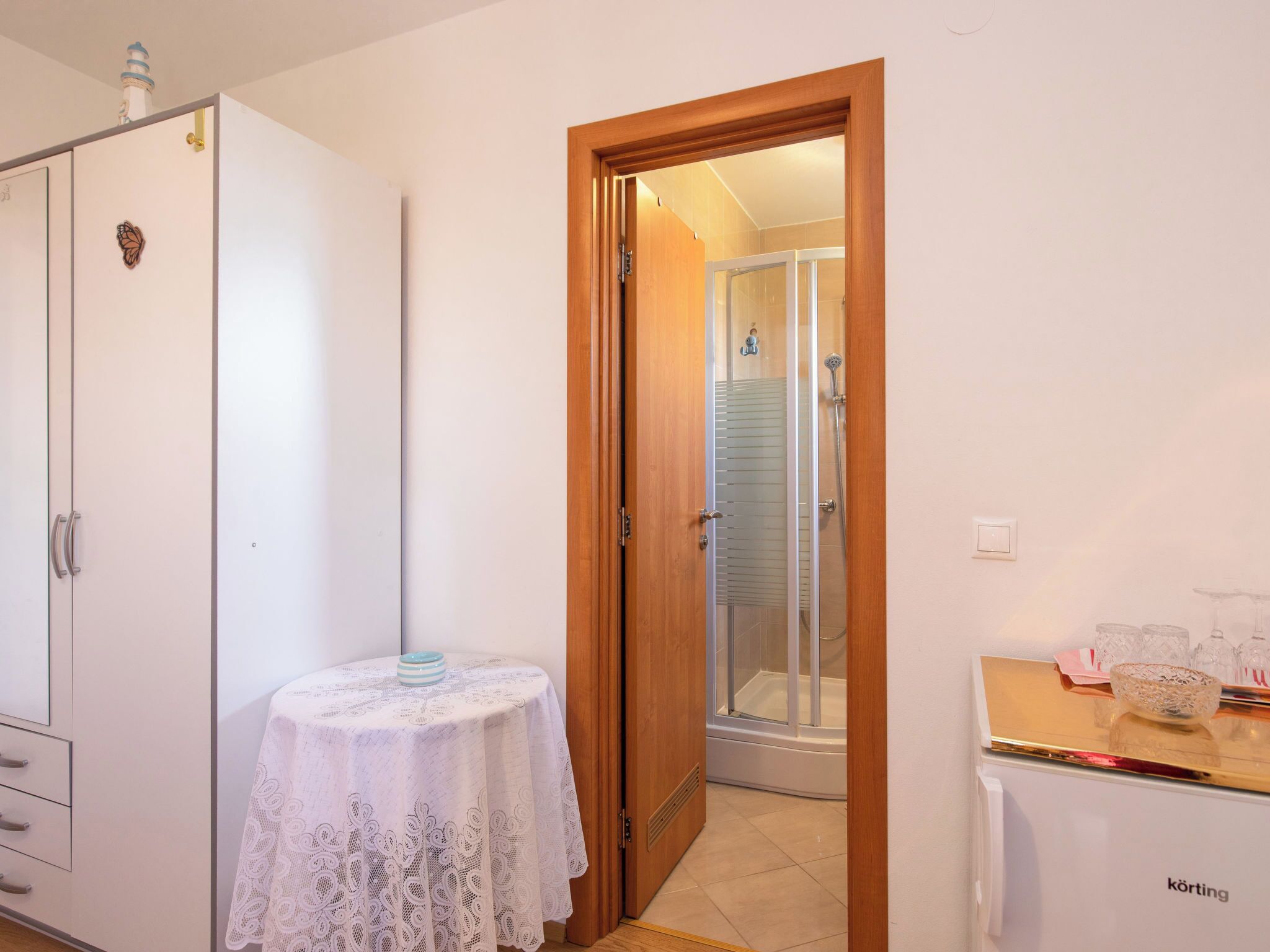 Une chambre dotée d’une armoire, d’une petite table recouverte d’une nappe en dentelle, d’un espace douche et d’un réfrigérateur.