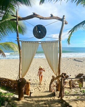 A beach scene with a wooden structure, palm trees, and a person walking towards the ocean. - Bohemia Beach Tayrona (Santa Marta)