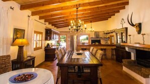 Un comedor con una larga mesa, sillas, una araña, una chimenea y una cocina al fondo.