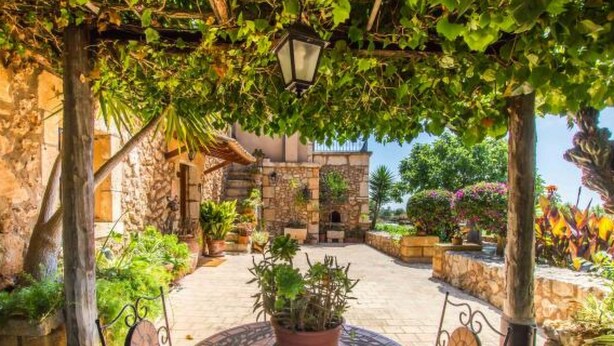 Un patio con una mesa redonda, plantas en macetas, muros de piedra y una pérgola con un farol.