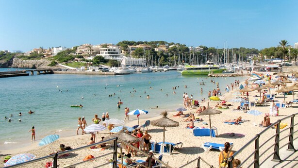 Una platja amb nombroses gandules i ombrel·les, una marina amb vaixells, i un poble al fons.