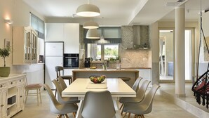 Une cuisine moderne dotée d’un îlot central, d’une table à manger avec des chaises, de luminaires suspendus et offrant une vue sur l’extérieur.