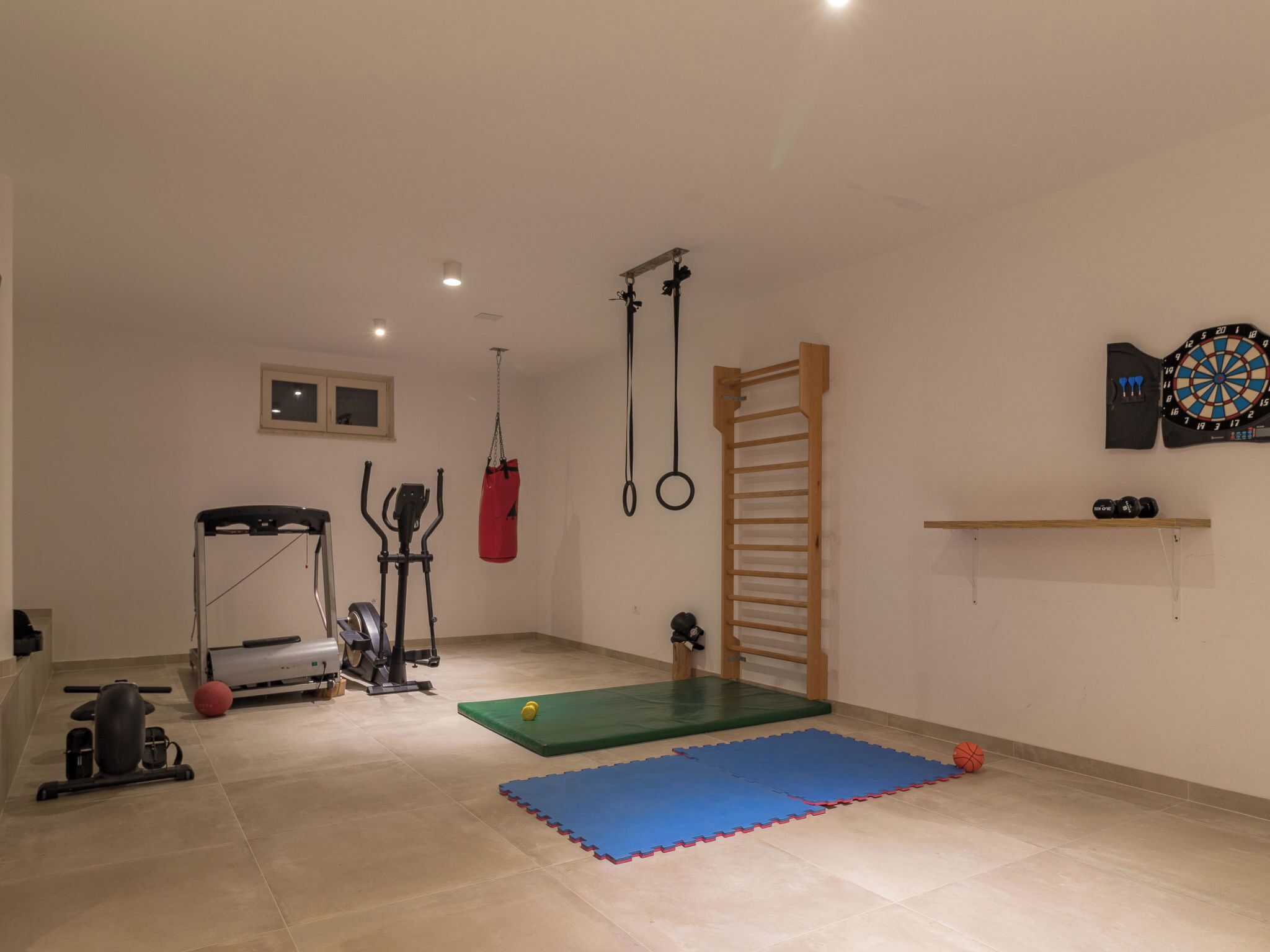 Een sportschool thuis met fitnessapparatuur, een pull-up stang, een groene fitnessmat en een dartbord.