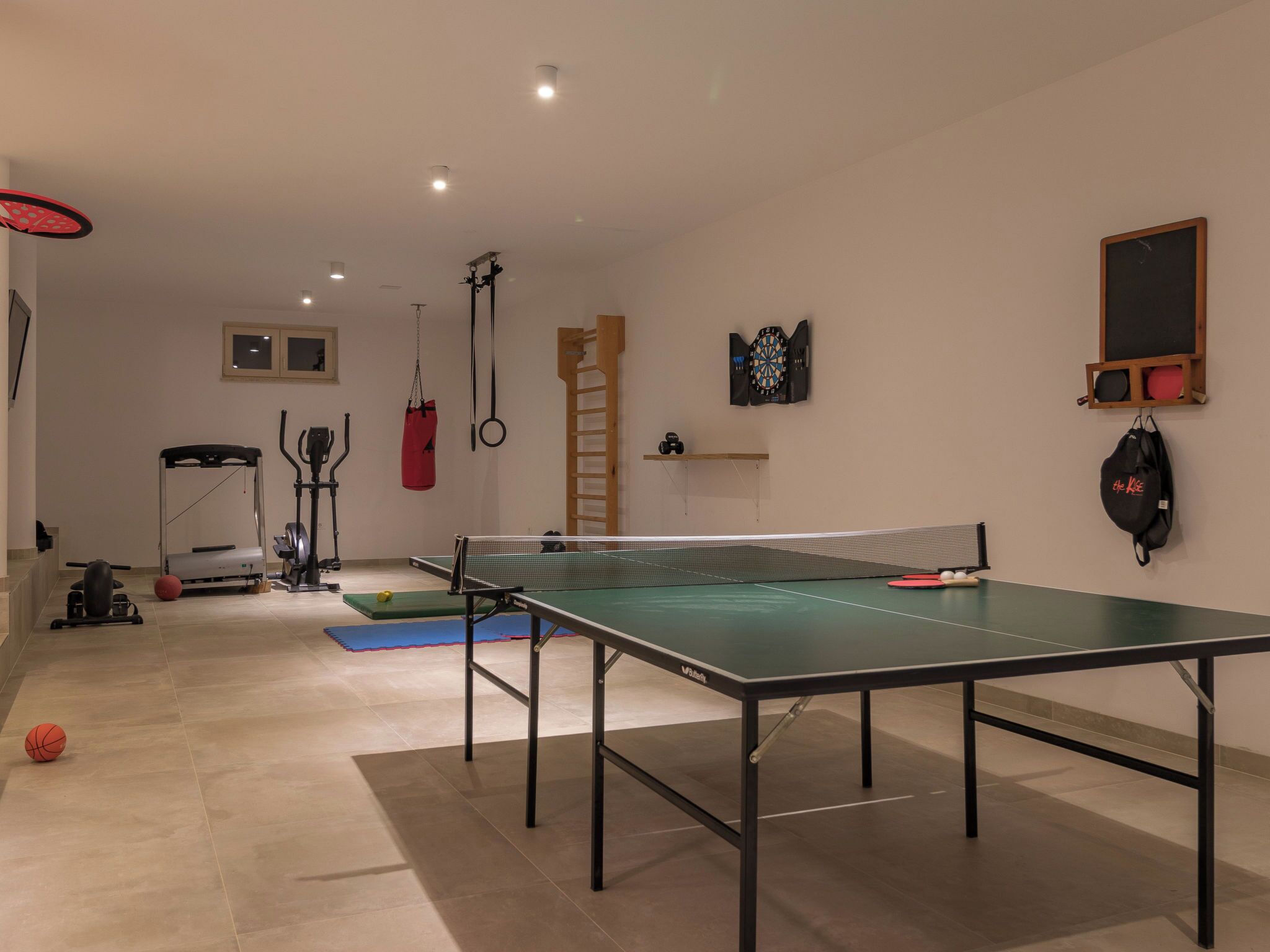Een kamer met een tafeltennistafel, sportapparatuur en een dartbord.