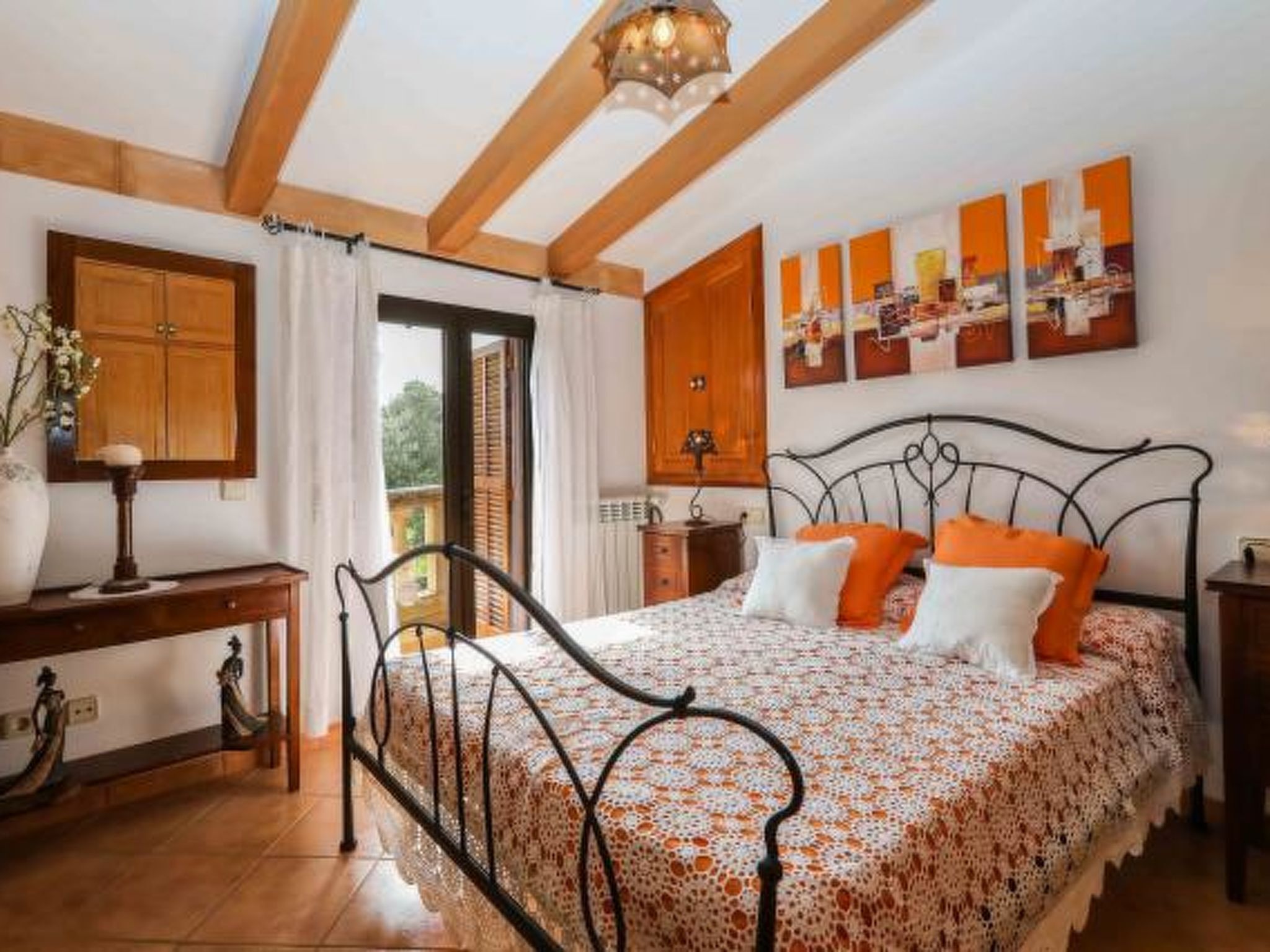 A bedroom with a bed, wooden ceiling beams, a desk, and a window with curtains.