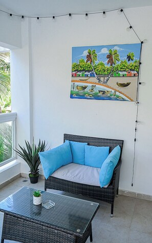 Une pi èce avec un canapé en osier, une table en verre, une plante en pot et un tableau représentant une scène de plage.