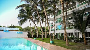 En swimmingpool med fliseterrasse, omgivet af palmer og en moderne bygning med altaner.