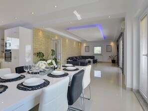 A modern dining area with a long table set for a meal, a kitchen area with an oven, and a living space with a sofa. - Villa Zeus by Interhome (Bokordici)