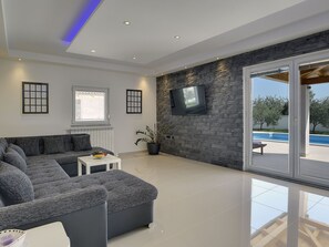 A modern living room with a sectional sofa, a flat-screen TV mounted on a stone wall, and a sliding glass door leading to an outdoor pool area. - Villa Zeus by Interhome (Bokordici)