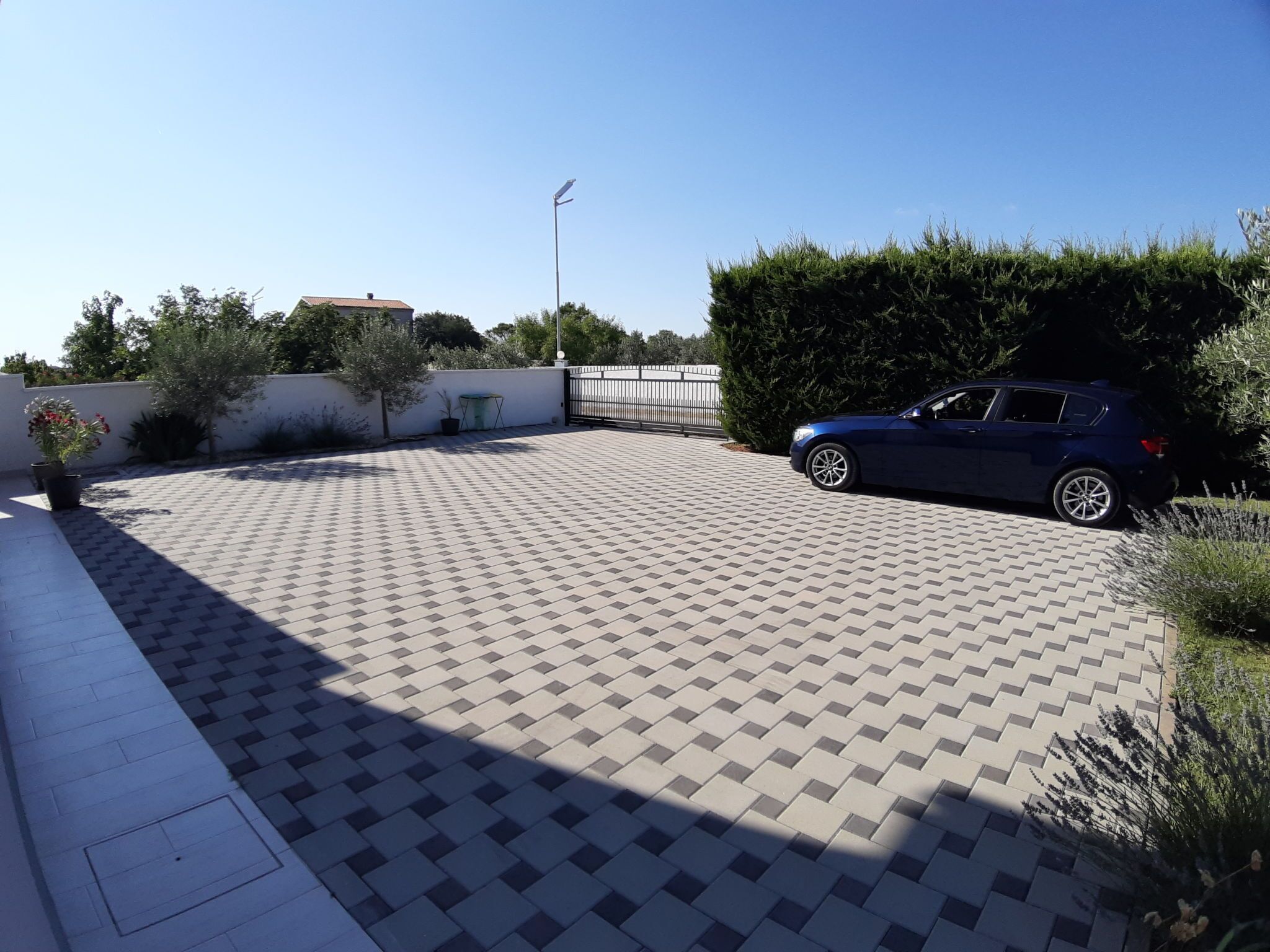 Een verhard buitengebied met een geparkeerde auto, omringd door groen en een witte muur.