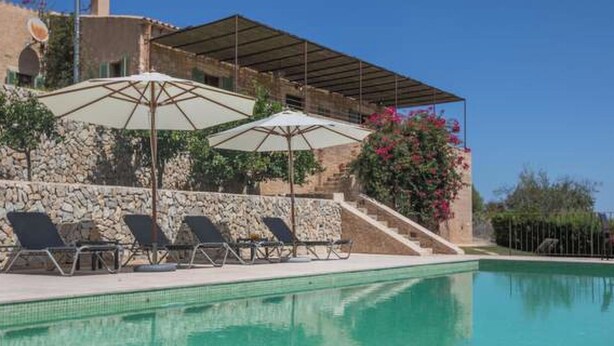 A swimming pool with lounge chairs and umbrellas, a stone wall, and a building with a sloped roof. - Vacation home Calma by Interhome (Sant Llorenç des Cardassar)