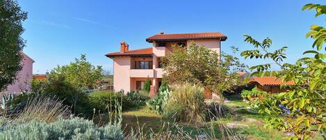A two-story house with a red-tiled roof, surrounded by lush greenery and a clear blue sky.