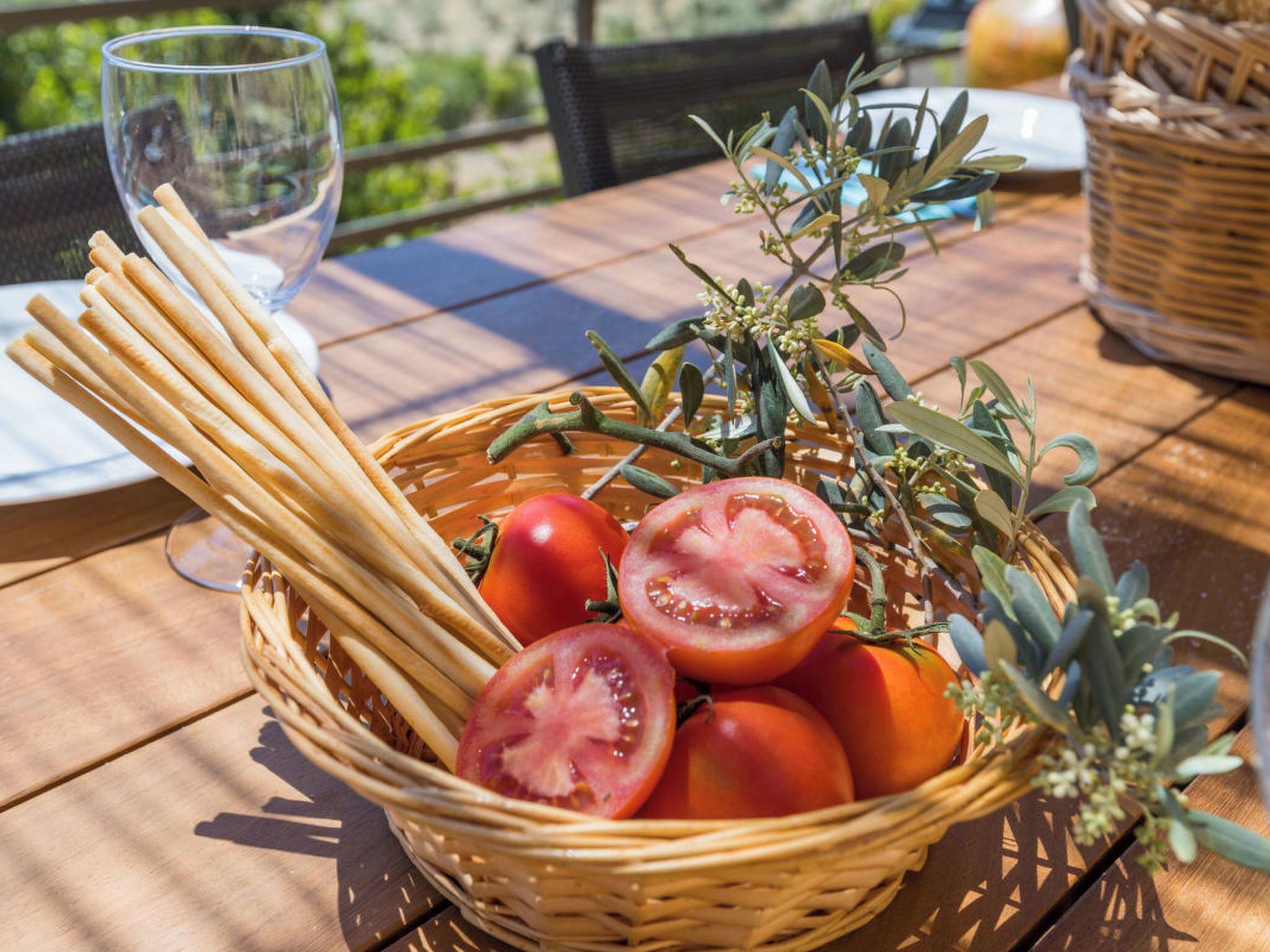 Un panier en osier rempli de tomates, un verre d’eau et un verre de vin sur une table à l’extérieur.