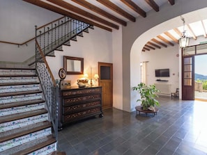 A staircase with decorative glass panels, a wooden dresser with drawers, a mirror, a lamp, and a television in a room with a tiled floor and an arched doorway leading outside. - Caballeria 16 Familiar by Interhome (Bunyola)