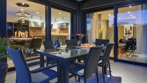 Un comedor moderno con una mesa de vidrio y sillas de mimbre, una cocina al fondo y vistas a través de grandes puertas de cristal.