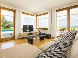 Ein modernes Wohnzimmer mit großem Fernseher, bequemen Sitzmöglichkeiten und Blick auf einen Swimmingpool.