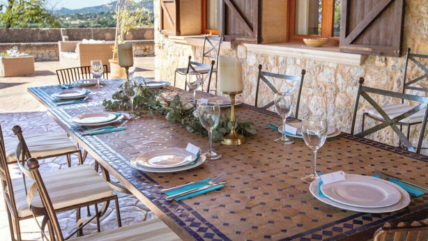 An outdoor dining area with a stone wall, wooden shutters, and a table set for six with chairs. - Maribel by Interhome (Inca)