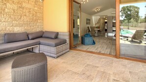 A modern living area with a stone wall, a grey sectional sofa, a wicker ottoman, and a glass sliding door leading to a dining area. - Villa Markulin by Interhome (Bokordici)