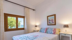 Una camera da letto con un letto, due comodini, una finestra con tende e una vista sul mare, oltre a un quadro appeso al muro.
