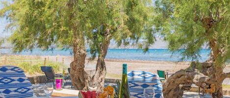 A table set for two with plates, cutlery, and a bottle of wine, overlooking a beach with trees and the sea.