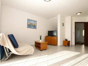 Ein Wohnzimmer mit Liegekorb, Fernseher auf einem Holzkasten, einem kleinen Tisch mit Pflanze und einer Tür zu einem weiteren Raum.