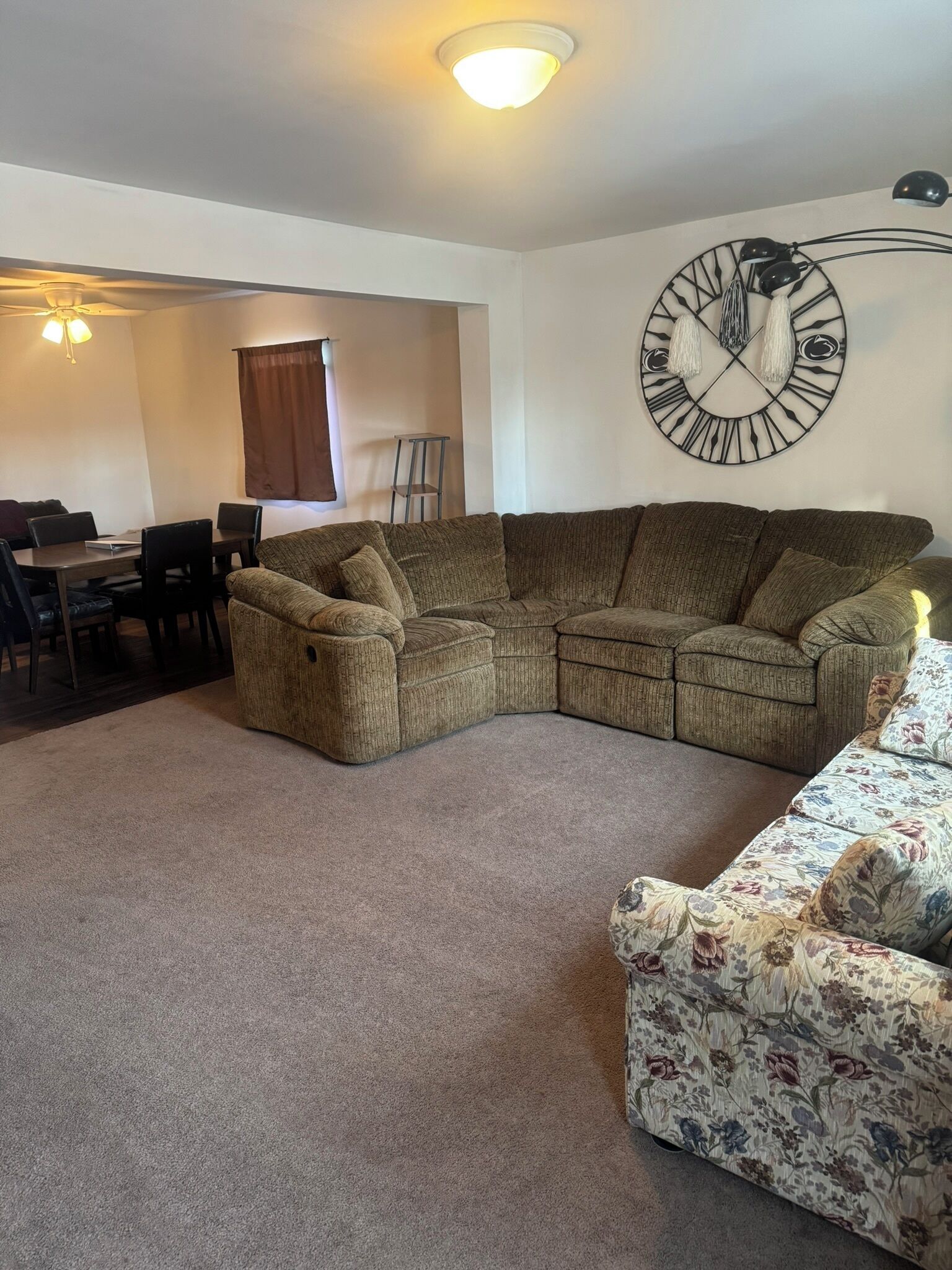 A living room with a sectional sofa, a floral armchair, a dining table with chairs, and a wall-mounted clock.