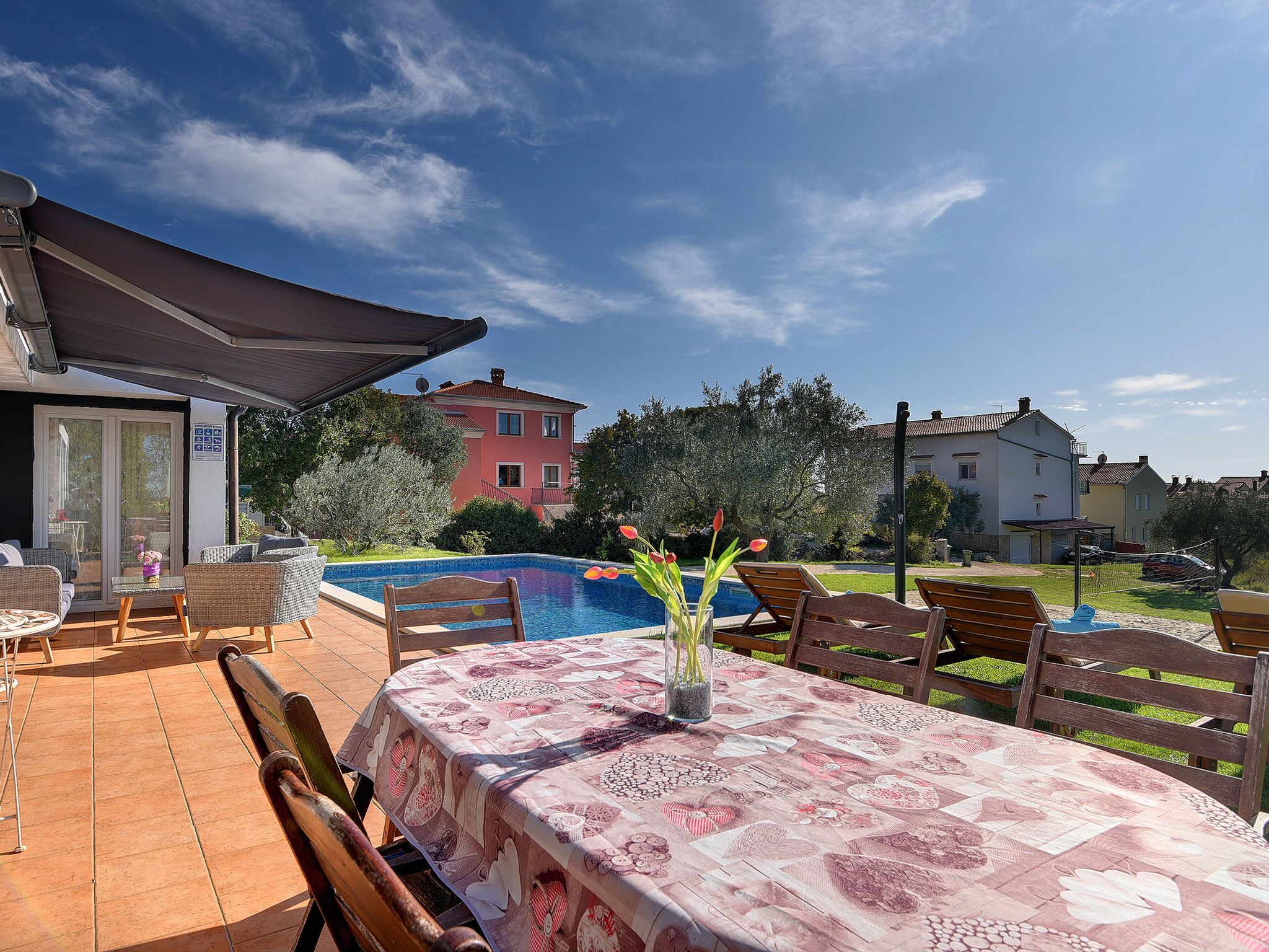 A covered patio with a pool, outdoor furniture, and a table with a patterned tablecloth.