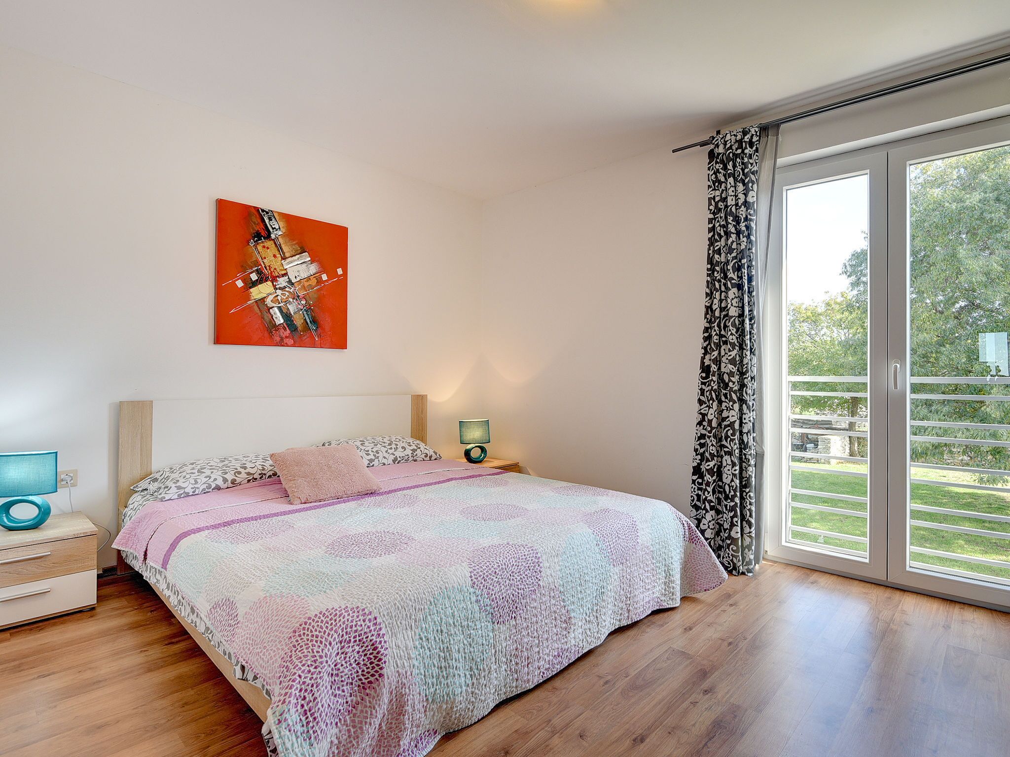 Een slaapkamer met een bed, nachtkastje, lamp, schilderij en een groot raam met gordijnen.