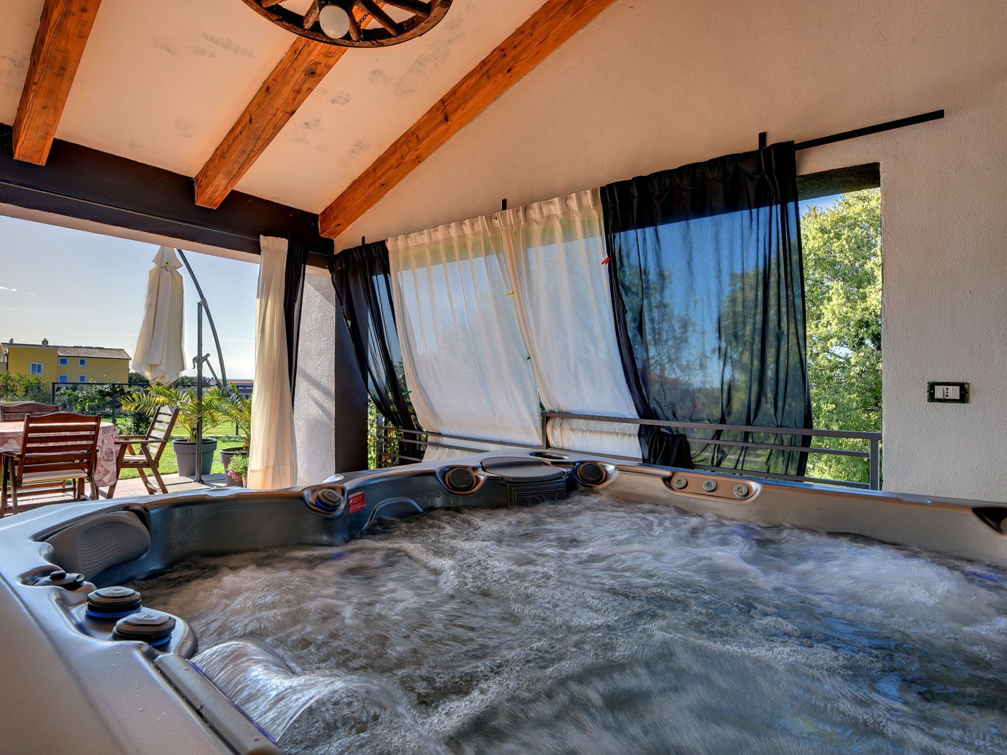 A hot tub in a room with wooden beams, large windows with curtains, and a view of greenery outside.