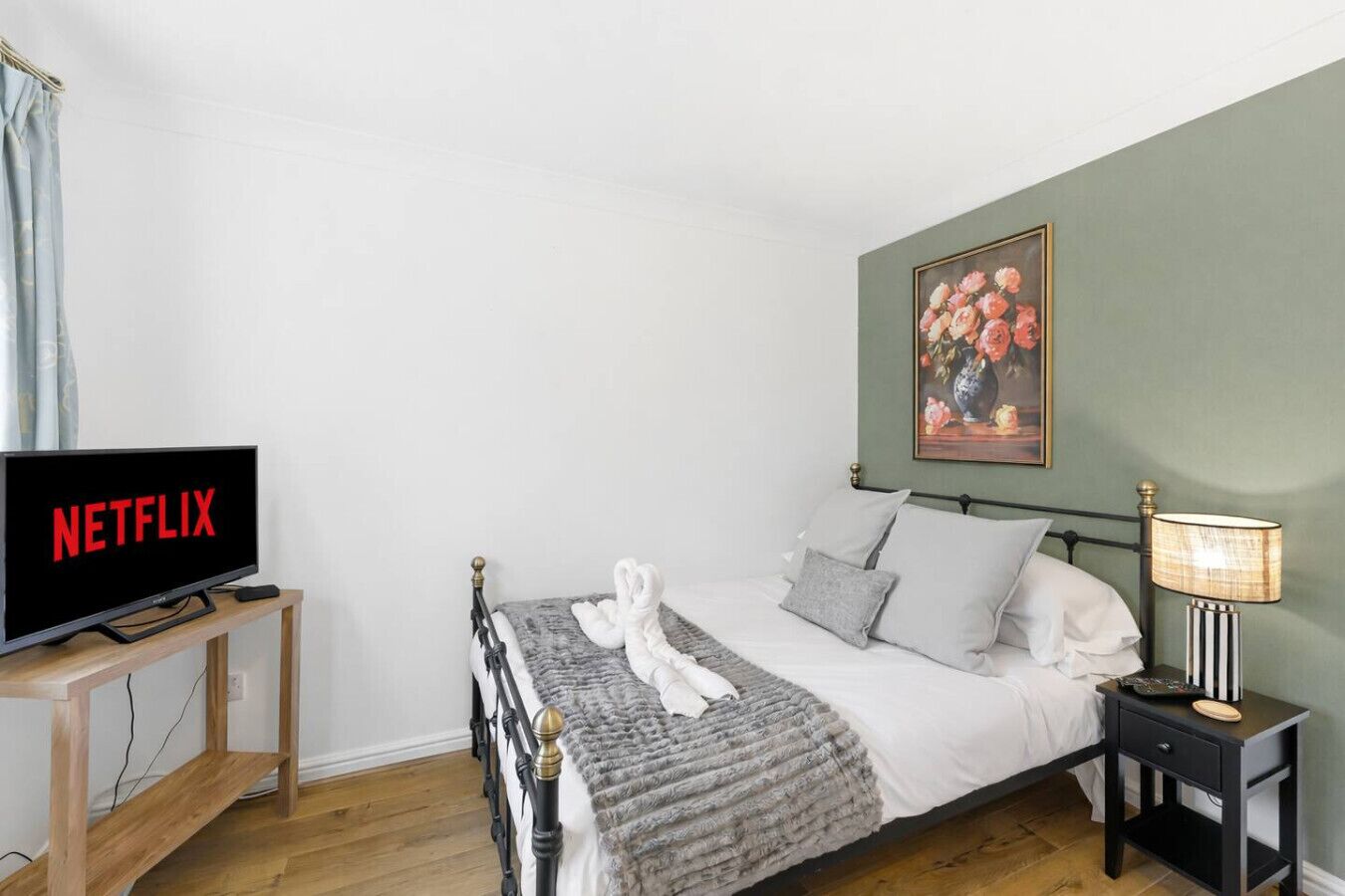 Un dormitorio ordenado con una cama, una mesita de noche con lámpara, un televisor mostrando 'NETFLIX' y un cuadro en la pared.