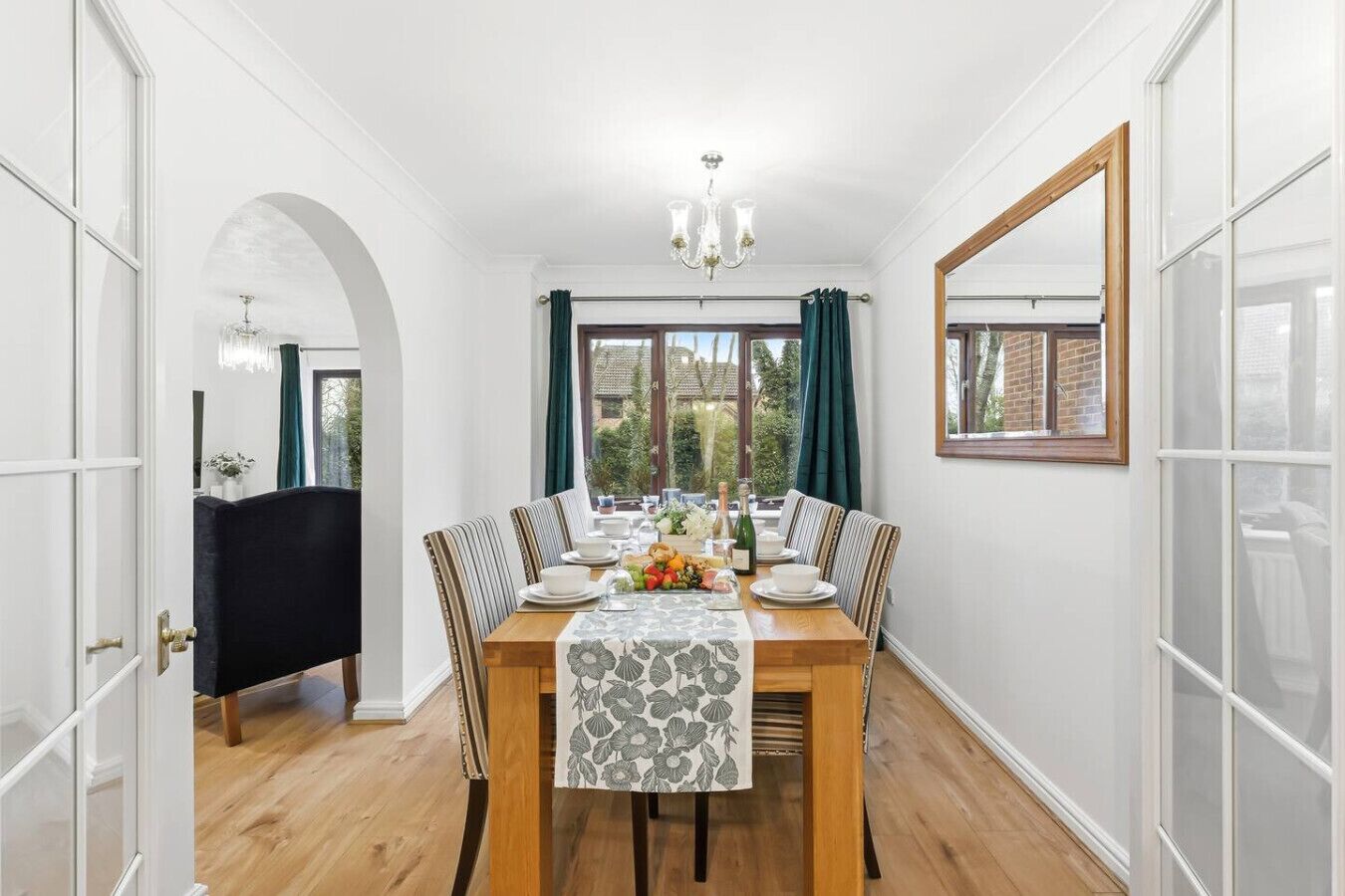 A dining room with a wooden table set for four, surrounded by chairs with striped cushions. A chandelier hangs from the ceiling, and a window with curtains is visible.