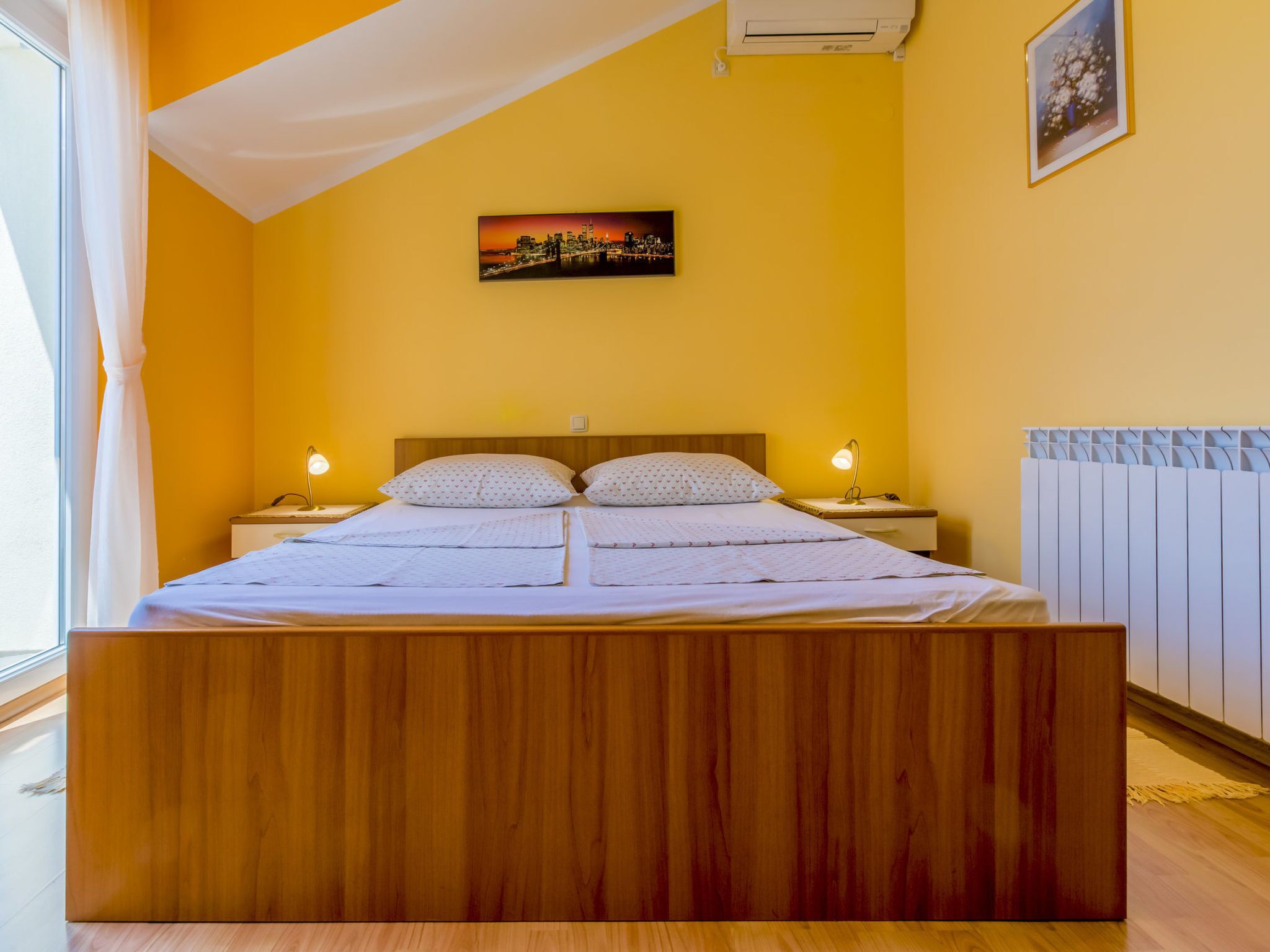 A bedroom with a wooden bed, two bedside tables with lamps, a cityscape painting on the wall, and a radiator.