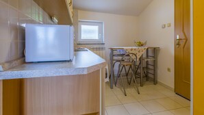 A compact kitchen with a refrigerator, a dining table with chairs, and a window. - BOZIDAR 1 zuti / HR-10438-01 by Interhome (Selce)