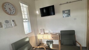 Un salon comprenant un bureau en bois, deux chaises, un téléviseur à écran plat, une horloge et une fenêtre avec des stores.