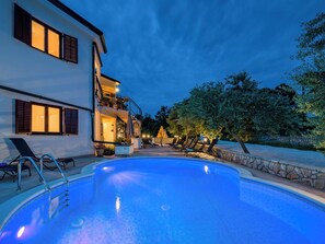 Vue sur une piscine, avec en arrière-plan un immeuble de deux étages doté d’un balcon, de chaises longues et d’un mur en pierre.