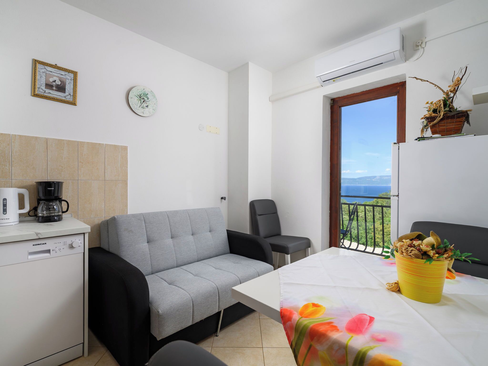A compact living space with a kitchenette, a grey sofa, a dining table with a colorful tablecloth, and a balcony with a view.