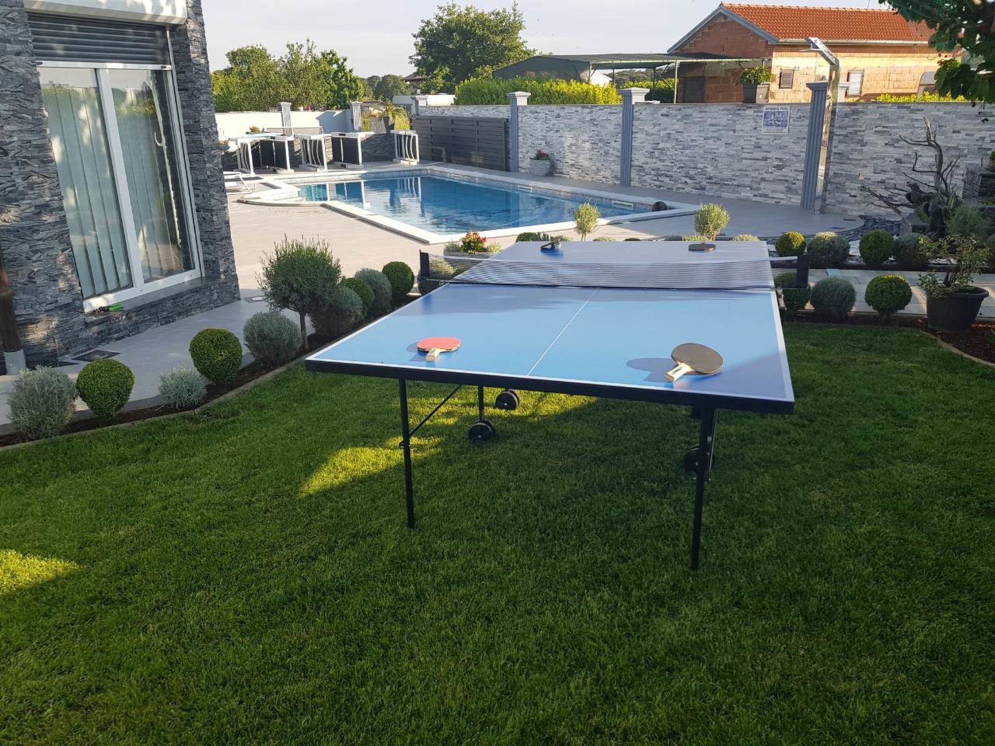 Een tafeltennistafel in een achtertuin met een zwembad en stenen muren.