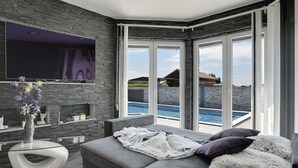 Ett modernt vardagsrum med en grå soffa, en platt-TV, en öppen spis och utsikt över en swimmingpool.