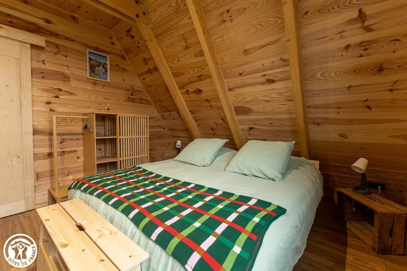 Un dormitorio acogedor en el ático, con techo inclinado de madera, cama con manta escocesa, mesita de noche y banco de madera.