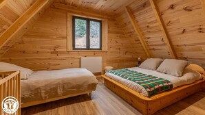 A cozy attic bedroom with a wooden sloped ceiling, a large window, a bed with a colorful blanket, and a small bedside table. - Le Chalet du Pastoureau (BESSE ET SAINT ANASTAISE)